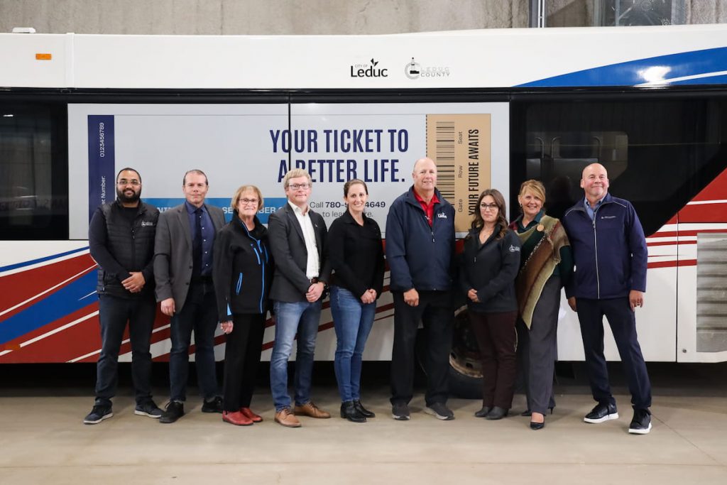 A group of people stand in front of a bus with a sign reading "Your Ticket to a Better Life" on its side.