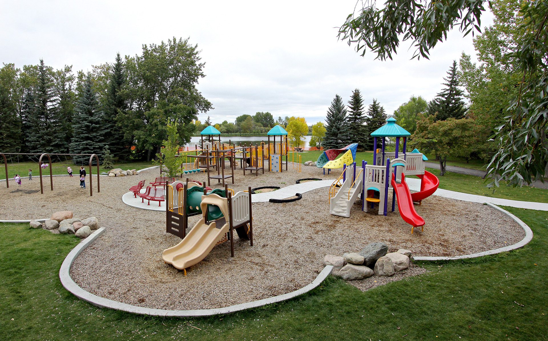 http://Children's%20playground%20with%20slides,%20climbing%20structures,%20and%20swings%20surrounded%20by%20trees%20and%20grass.%20A%20few%20kids%20are%20playing%20in%20the%20area.
