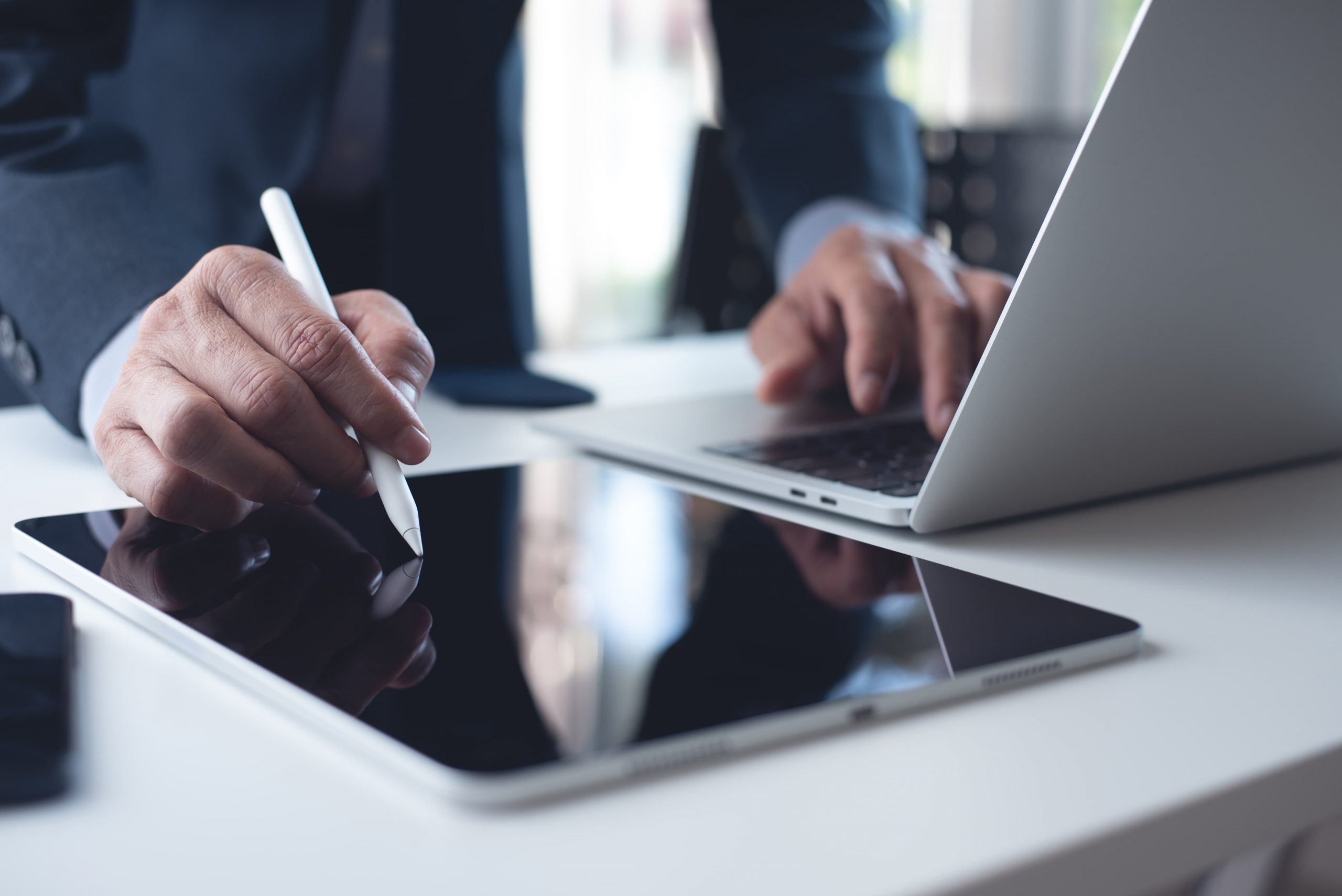 A person uses a stylus on a tablet while working on a laptop at a desk.