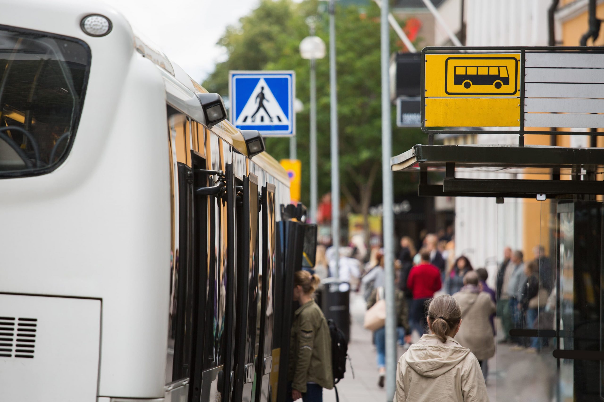 http://Bus%20stop%20with%20a%20yellow%20sign,%20a%20waiting%20bus,%20and%20several%20people%20boarding.%20Trees%20and%20a%20pedestrian%20crossing%20sign%20are%20visible%20in%20the%20background.