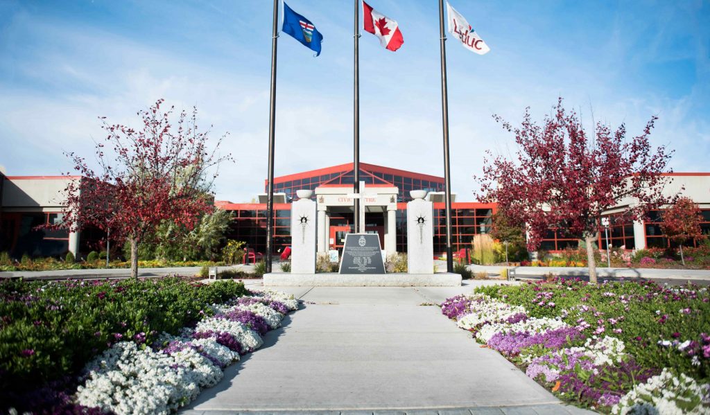 Pathway leads to a building with three flagpoles displaying Alberta, Canadian, and another flag. Flower beds with red, white, and purple flowers border the walkway.