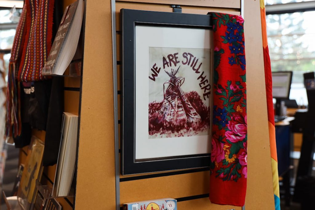 Framed art with a teepee and "We Are Still Here" text, displayed on a shelf with a vibrant floral fabric draped over the corner.