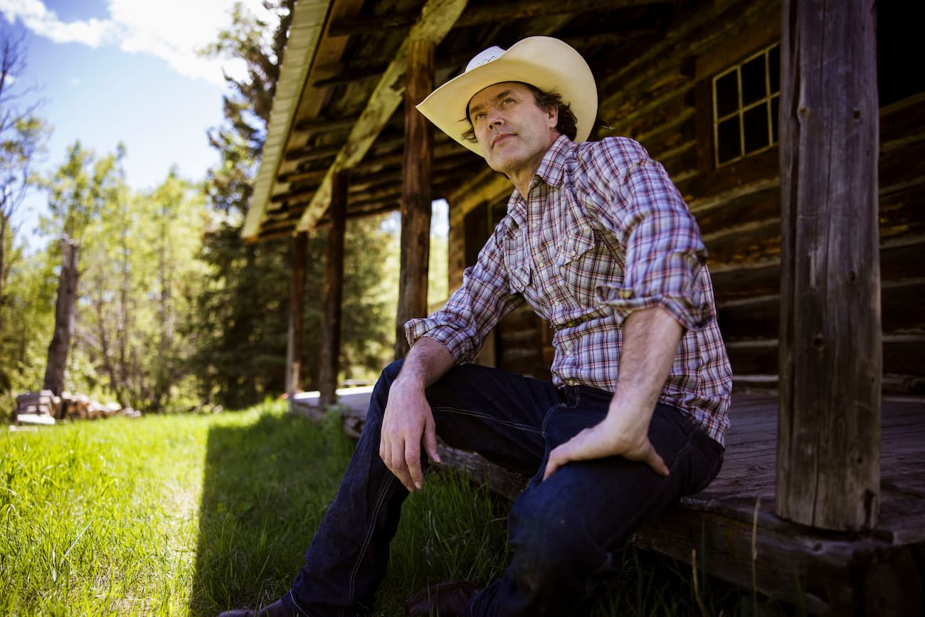 Country singer Corb Lund sitting outside a log cabin wearing a cowboy hat and plaid shirt