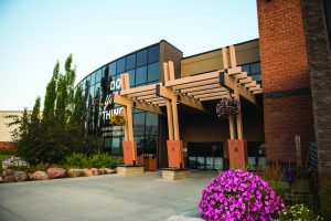 Modern building entrance with glass facade and wooden pergola. Pink flowers and greenery surround the walkway.