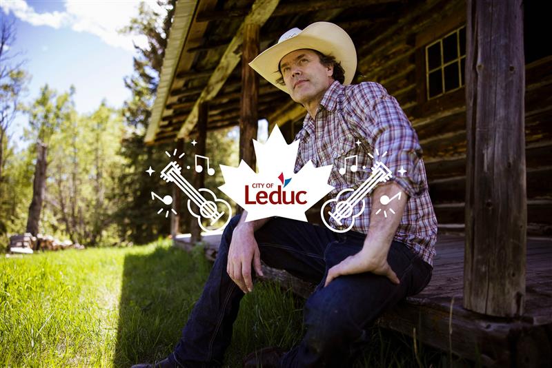 Country singer Corb Lund sitting outside a log cabin wearing a cowboy hat and plaid shirt