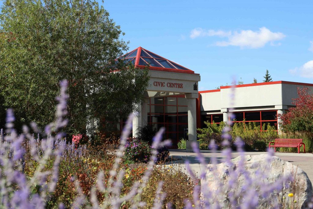 Civic Centre building with a red-trimmed roof, surrounded by trees and purple flowers under a clear blue sky.