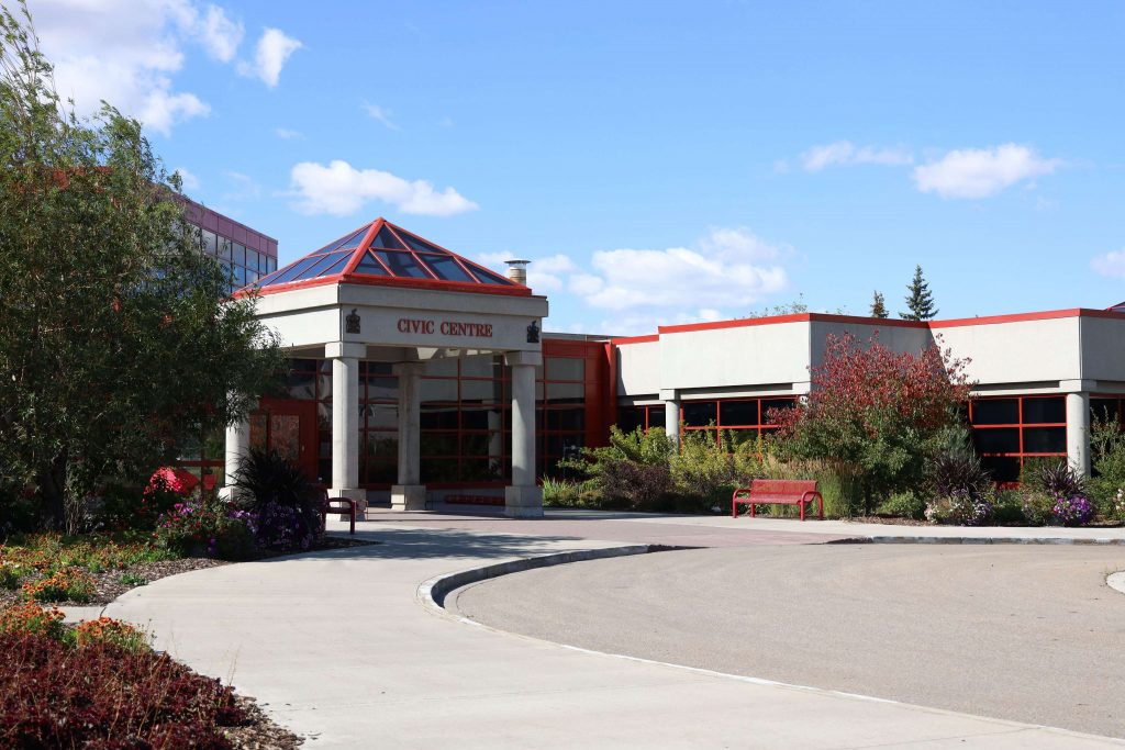 Entrance of a civic center with a glass pyramid roof, surrounded by trees and benches, under a clear blue sky.