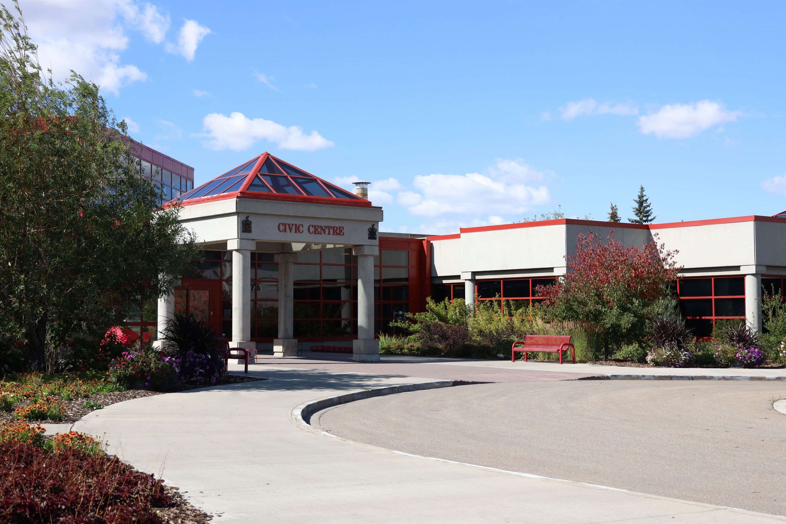Entrance of a civic center with a glass pyramid roof, surrounded by trees and benches, under a clear blue sky.