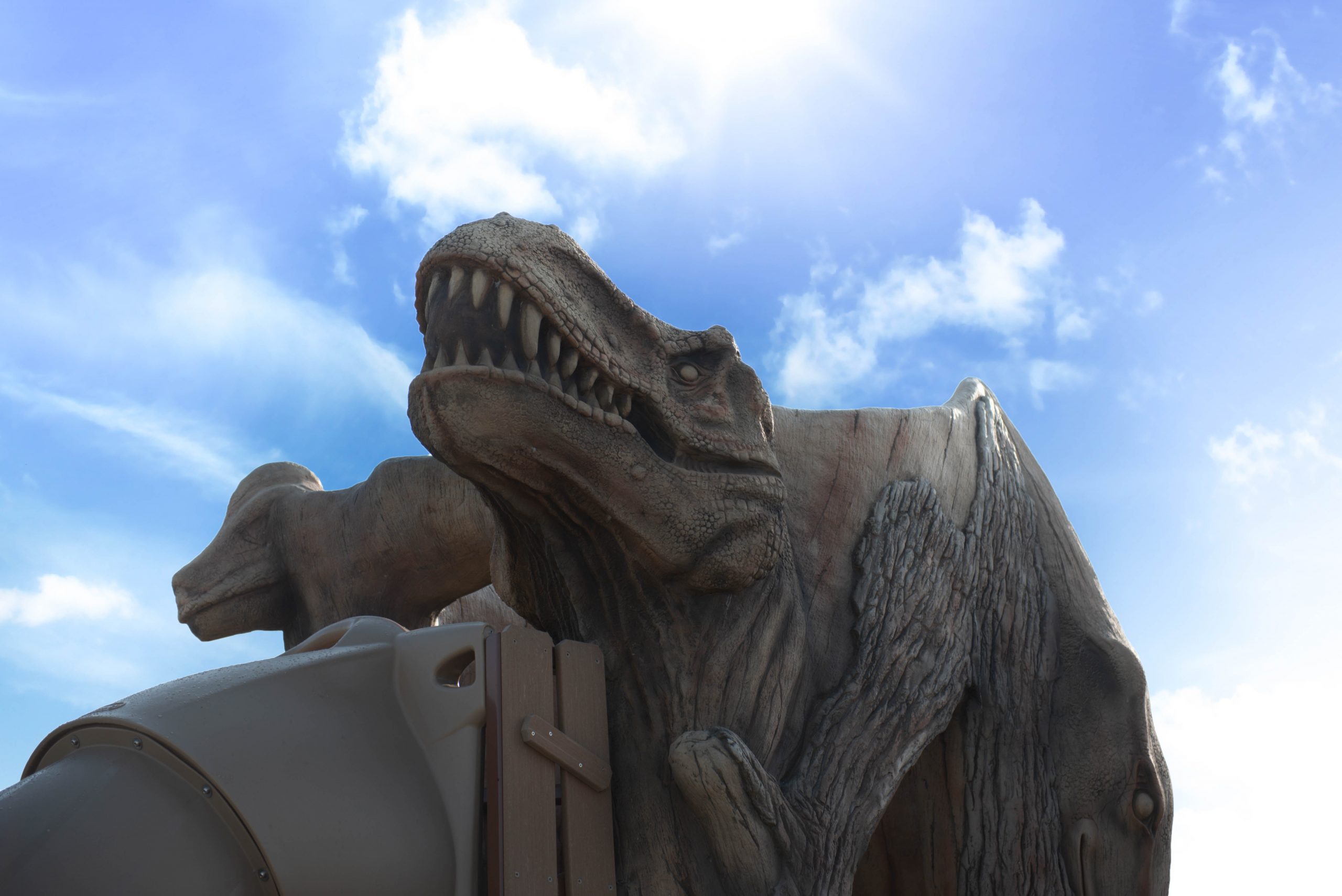 A large dinosaur sculpture with sharp teeth against a blue sky with clouds and bright sunlight.