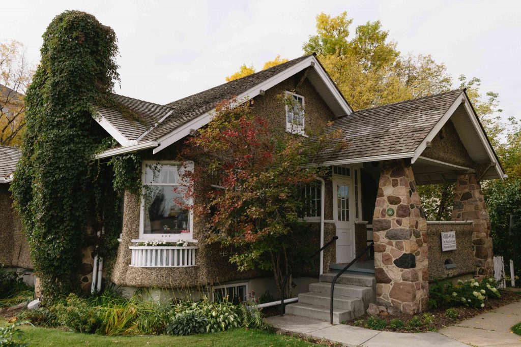 A stone and stucco house with a steep roof, chimney, and a front porch, surrounded by trees and shrubs.