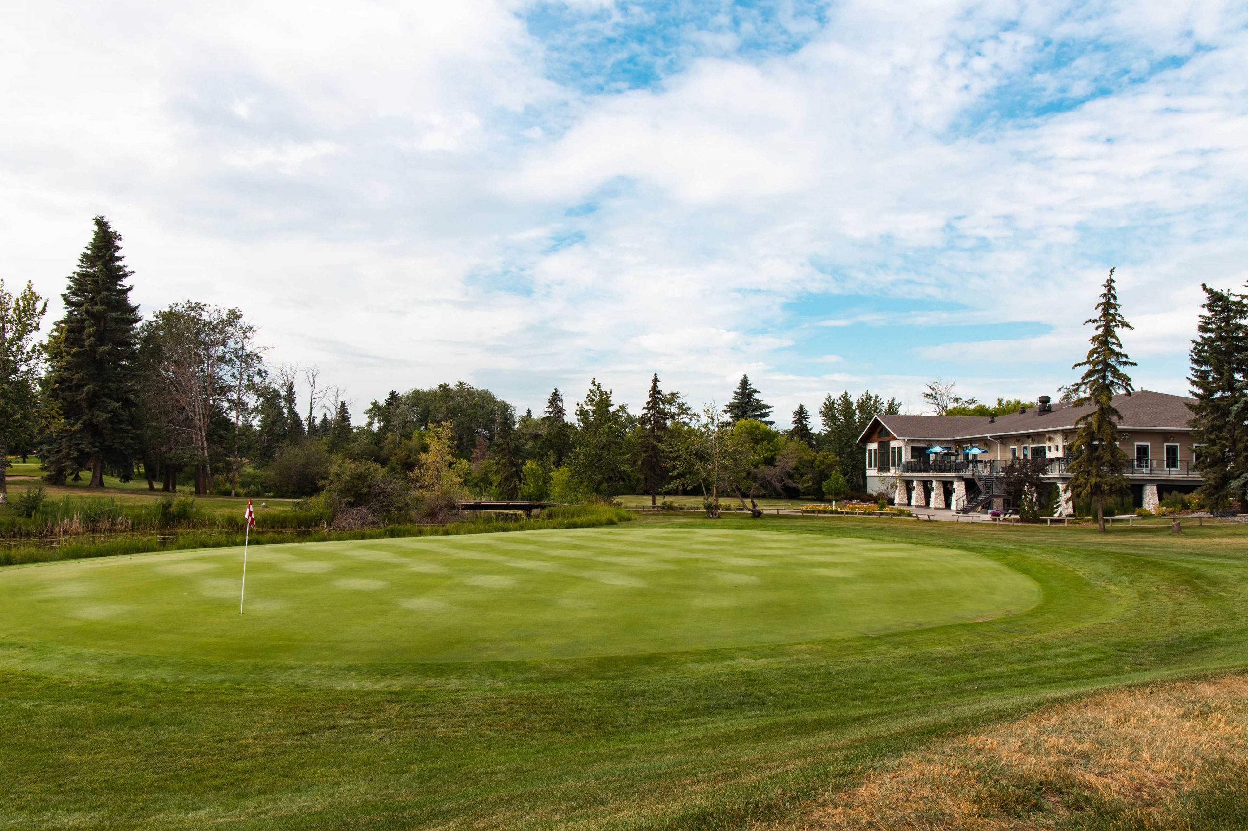 Golf course with a flag on the green, surrounded by trees and a clubhouse in the background under a partly cloudy sky.
