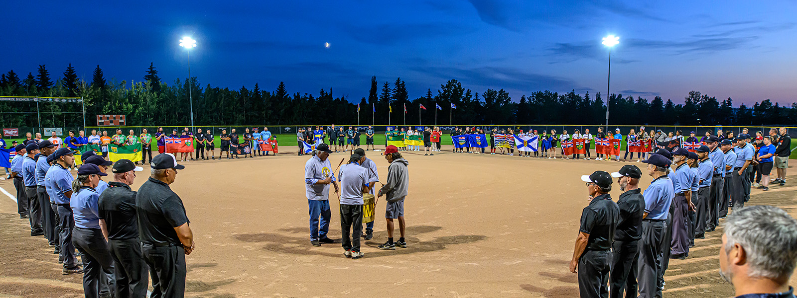 http://Teams%20and%20officials%20in%20uniforms%20gather%20on%20a%20softball%20field%20under%20stadium%20lights%20at%20dusk,%20forming%20a%20large%20circle.