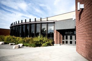 Modern building with large glass windows and a brown brick exterior, surrounded by greenery and rocks under a partly cloudy sky.