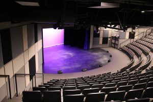 Empty auditorium with curved rows of gray seats facing a stage with purple lighting and a dark blue curtain.