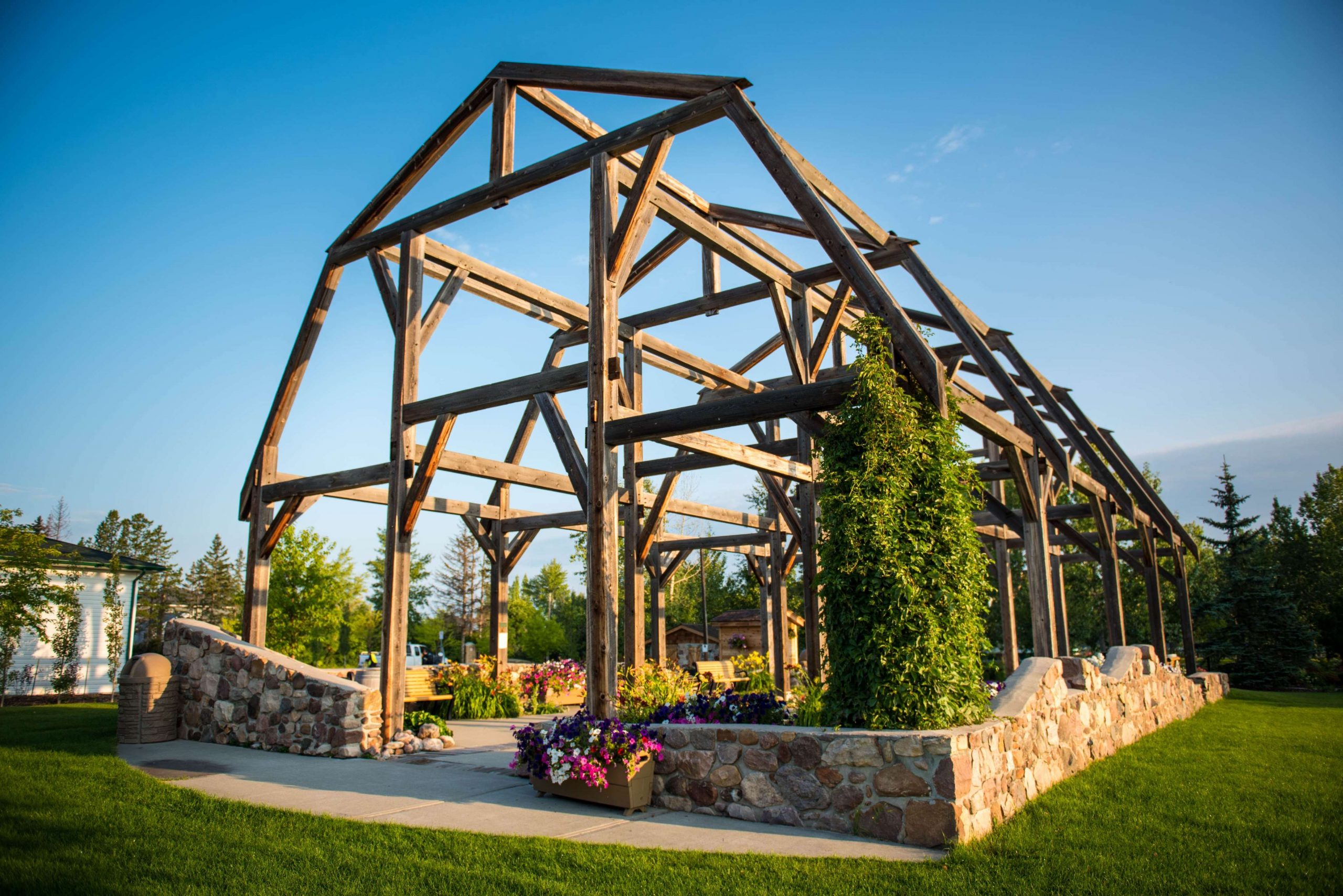 Wooden frame structure stands in a garden with stone walls and vibrant flowers, set against a clear blue sky.