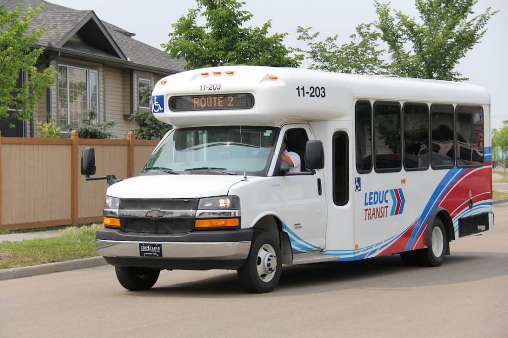 Small white and blue Leduc transit bus on Route 2 driving on a suburban street with houses and trees in the background.