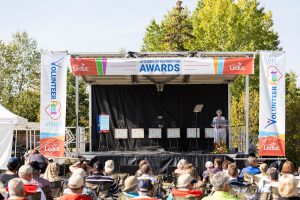 Outdoor stage with a speaker at a podium, audience seated, banners reading "Volunteer Fest" and "Awards" visible.
