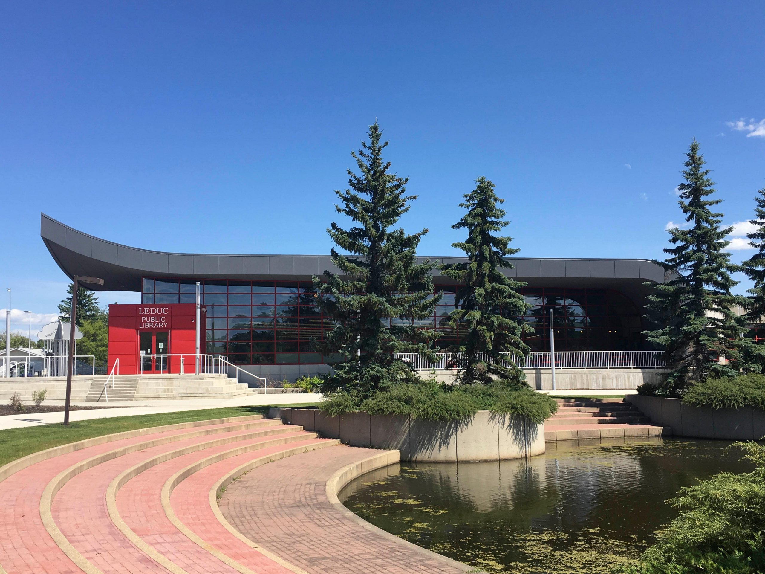 http://Modern%20library%20building%20with%20large%20windows,%20surrounded%20by%20pine%20trees%20and%20a%20curved%20brick%20pathway%20next%20to%20a%20pond%20under%20a%20clear%20blue%20sky.