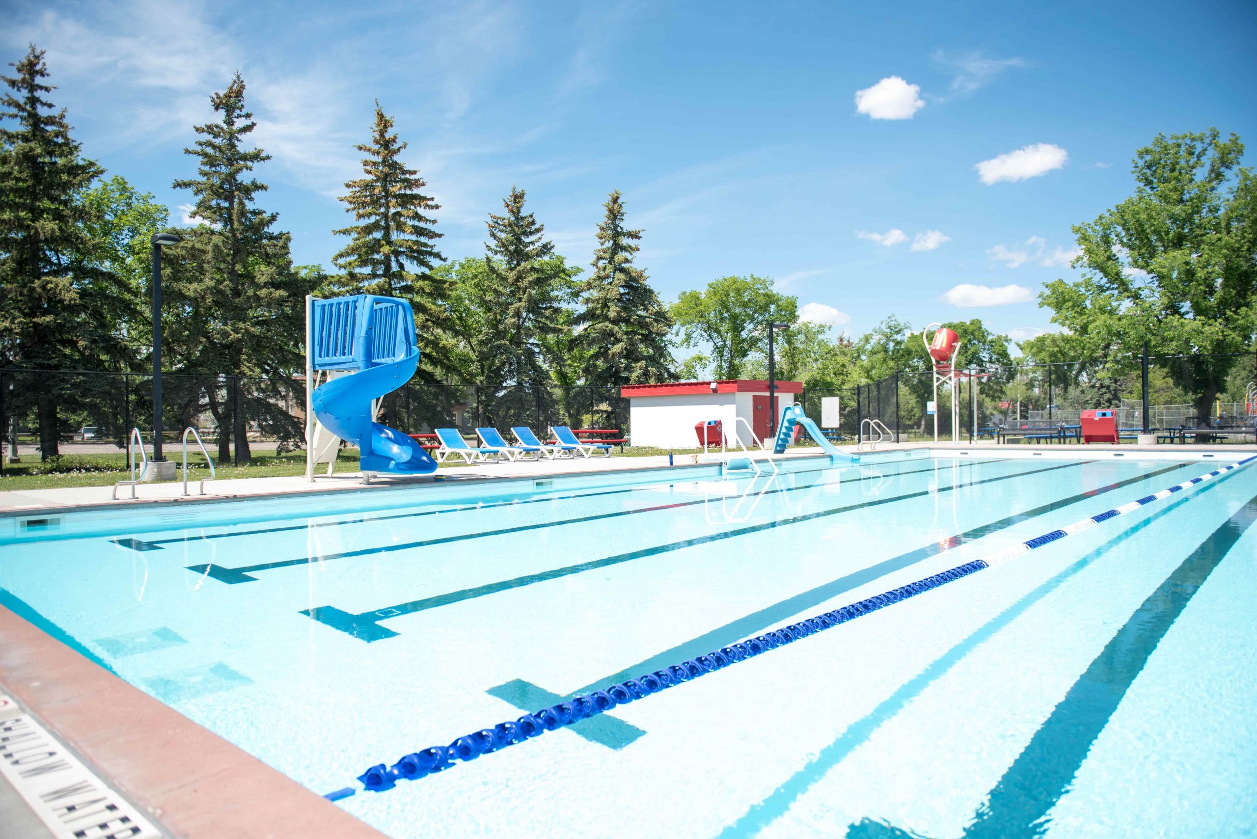 Outdoor swimming pool with a water slide, lounge chairs, and trees in the background on a sunny day.