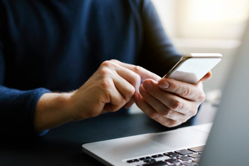 Person in dark sweater uses a smartphone with both hands, sitting in front of a laptop on a desk.
