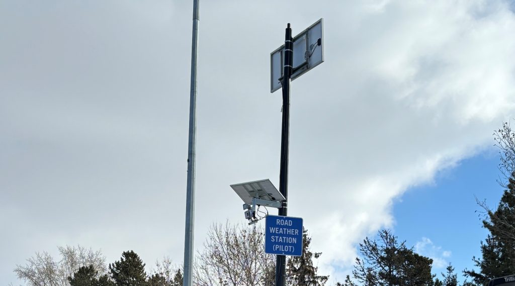 Weather station on a tall pole with attached solar panels and a pilot sign, set against a cloudy sky with trees below.