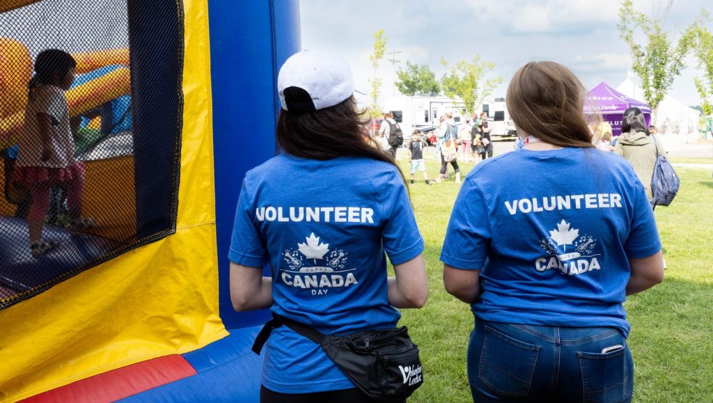 Two volunteers in blue "Canada Day" shirts stand near an inflatable structure at an outdoor event with people around.