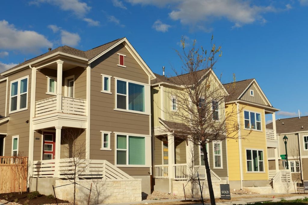 Row of modern two-story houses with balconies and colored facades under a blue sky. One house has a "sold" sign in front.