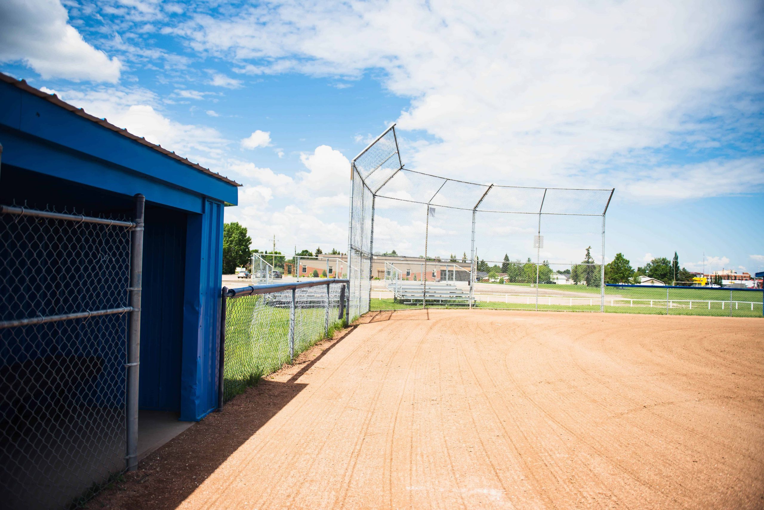 Empty baseball field with a blue dugout, chain-link fencing, and a grassy outfield under a partly cloudy sky.