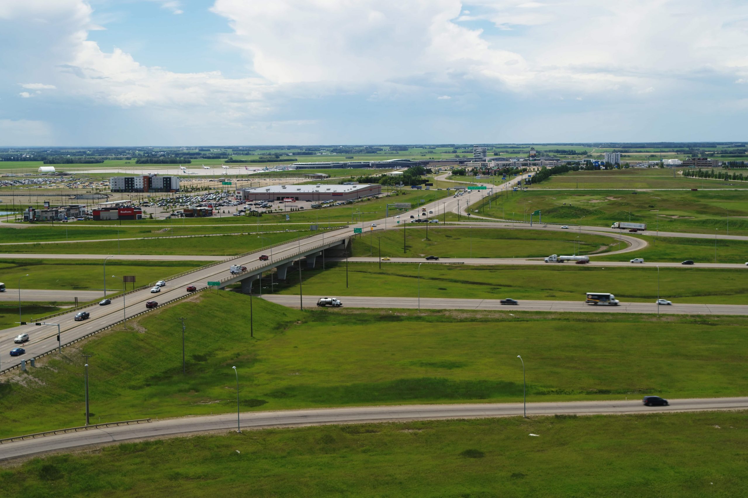 Aerial view of a highway interchange in a green landscape with scattered vehicles and buildings under a partly cloudy sky.