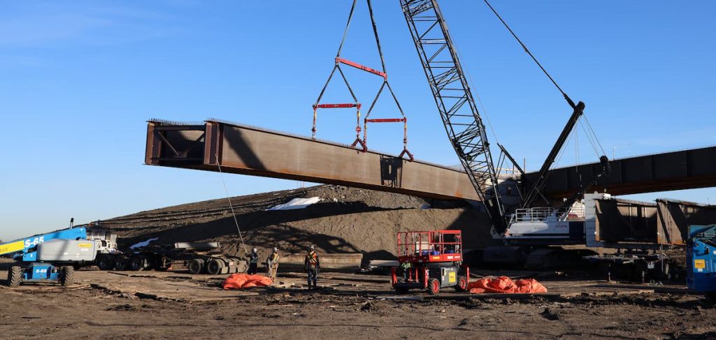 Crane lifting large steel beam at construction site with workers and equipment under clear sky.