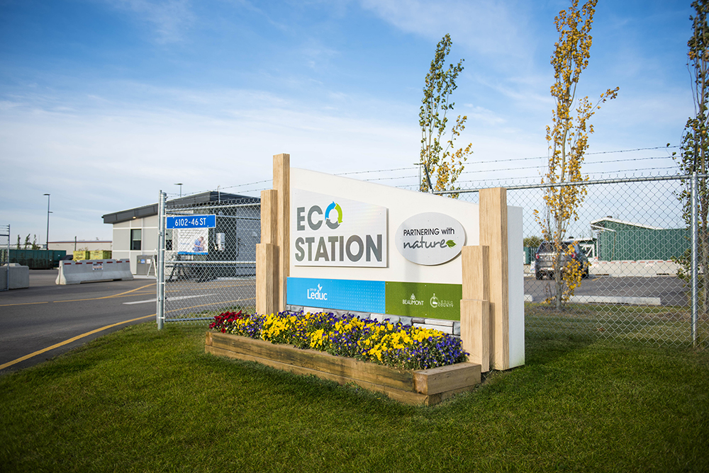 http://Eco%20Station%20sign%20with%20flowers%20in%20front,%20located%20on%20a%20grassy%20area%20near%20a%20fenced%20facility%20and%20a%20building%20in%20the%20background.