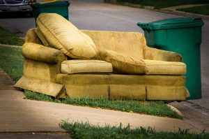 Old yellow couch and green trash bins on a sidewalk curb in a suburban neighborhood.