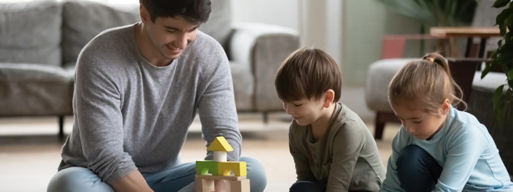 A man and two children build a wooden block tower together, sitting on the floor in a bright living room.