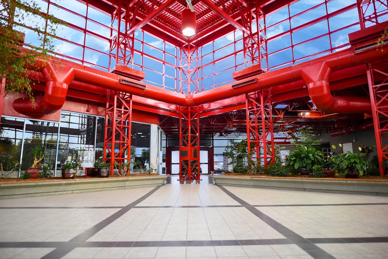 http://Spacious%20indoor%20area%20with%20red%20metal%20structures,%20large%20windows,%20ceiling%20lights,%20tile%20flooring,%20and%20plants%20along%20the%20walls.