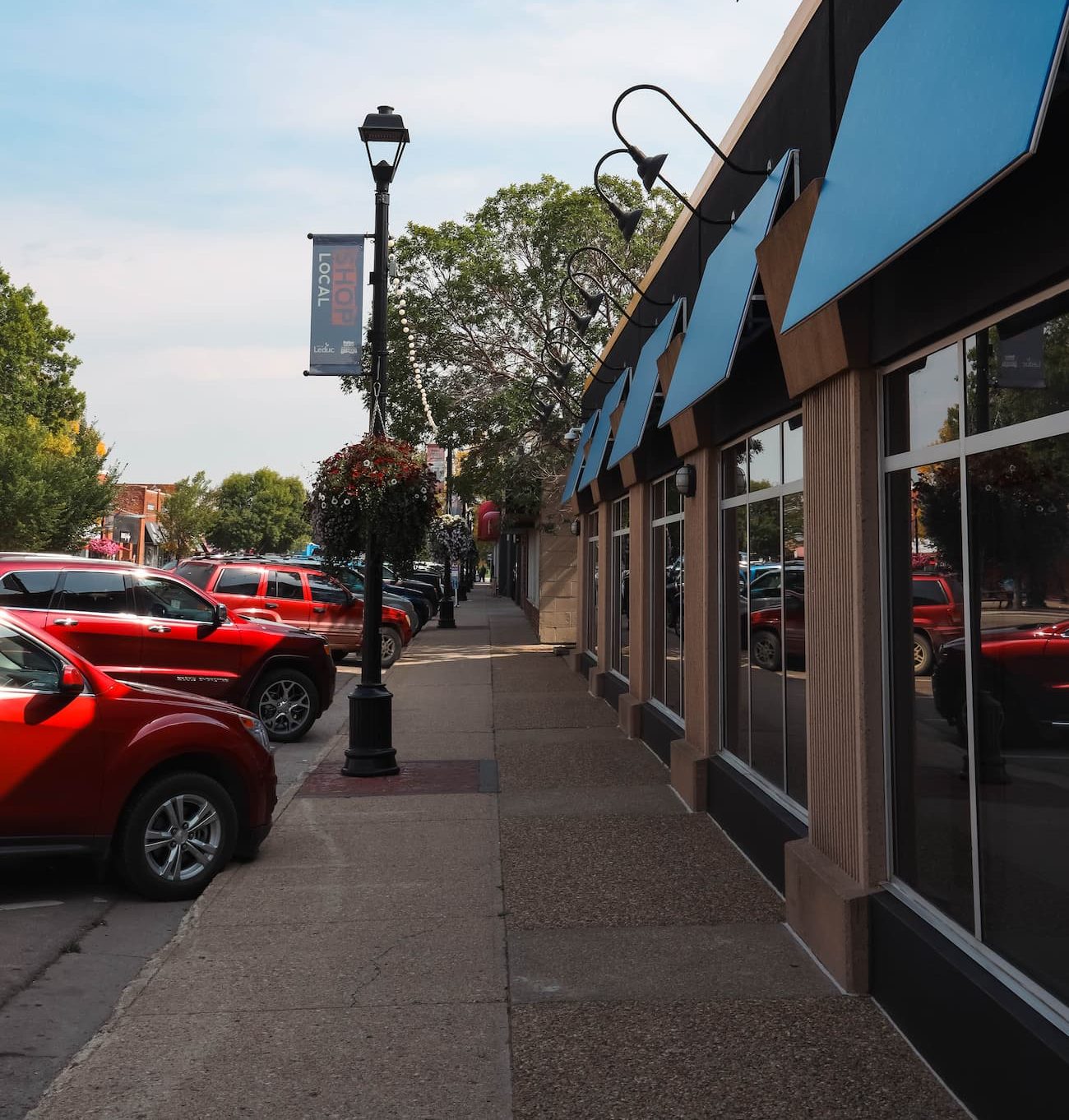 http://Street%20view%20of%20parked%20red%20cars,%20a%20row%20of%20blue%20awnings%20on%20a%20building,%20and%20a%20sidewalk%20with%20a%20lamp%20post%20and%20hanging%20flower%20basket.