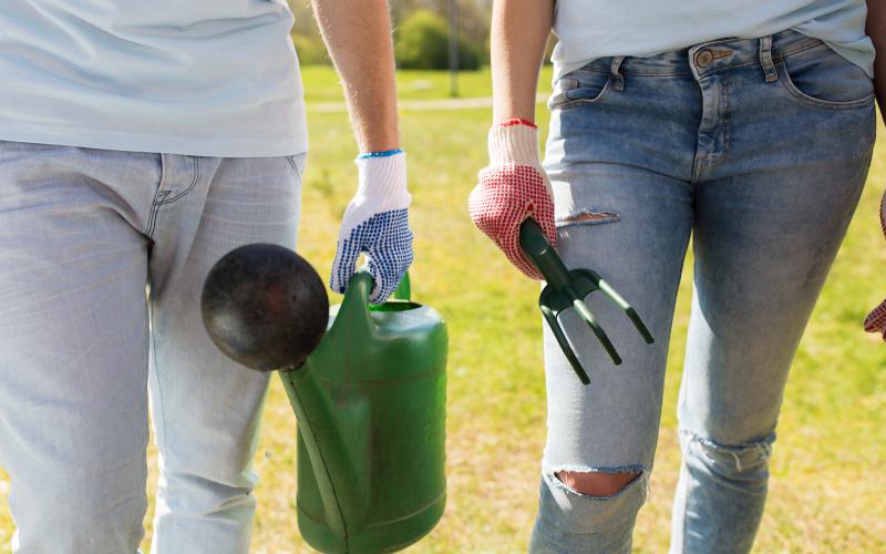 http://Two%20people%20holding%20gardening%20tools,%20including%20a%20watering%20can%20and%20fork,%20wearing%20gloves,%20standing%20on%20grassy%20ground.