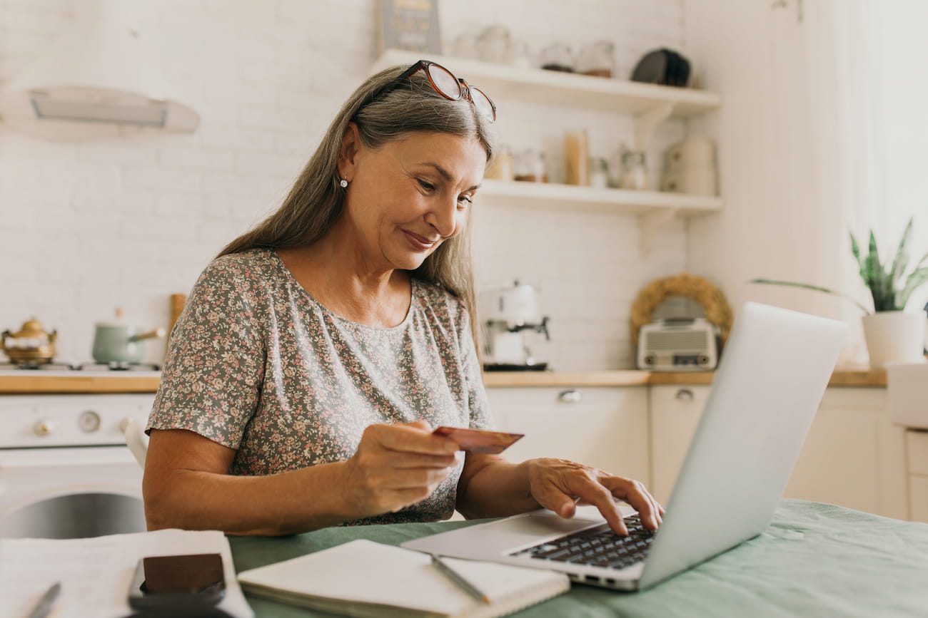 http://A%20woman%20with%20long%20gray%20hair%20holds%20a%20credit%20card%20while%20using%20a%20laptop%20in%20a%20kitchen.%20Notebook%20and%20pen%20beside%20her.