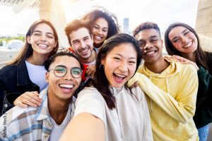 A group of seven diverse young adults smiling widely and taking a selfie outdoors during the day.