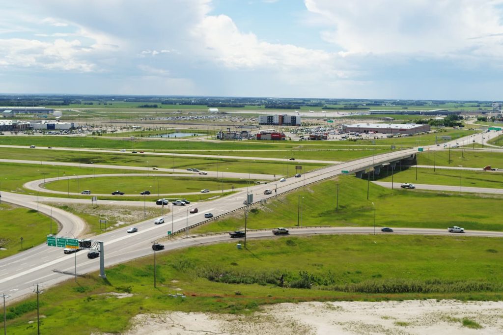 Aerial view of a highway interchange surrounded by green fields, with scattered buildings and a parking lot in the background.