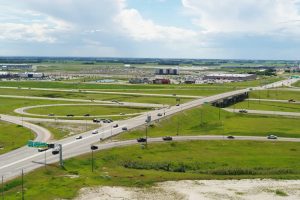 Aerial view of a highway interchange surrounded by green fields, with scattered buildings and a parking lot in the background.