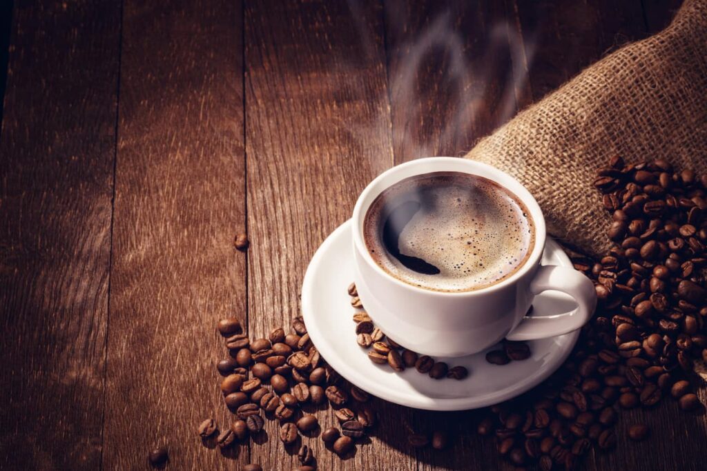 Hot cup of coffee with steam rising out of it surrounded by coffee beans on a wood plank table