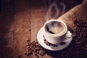 Hot cup of coffee with steam rising out of it surrounded by coffee beans on a wood plank table