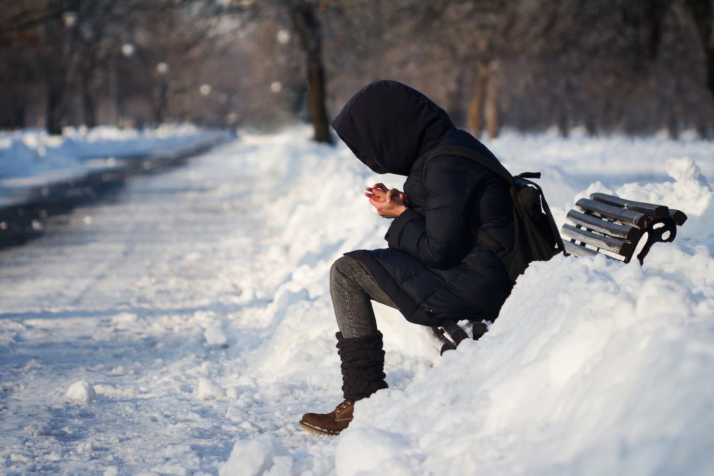 http://Person%20in%20winter%20coat%20sits%20on%20a%20snow-covered%20bench%20in%20a%20snowy%20park,%20looking%20at%20their%20hands.