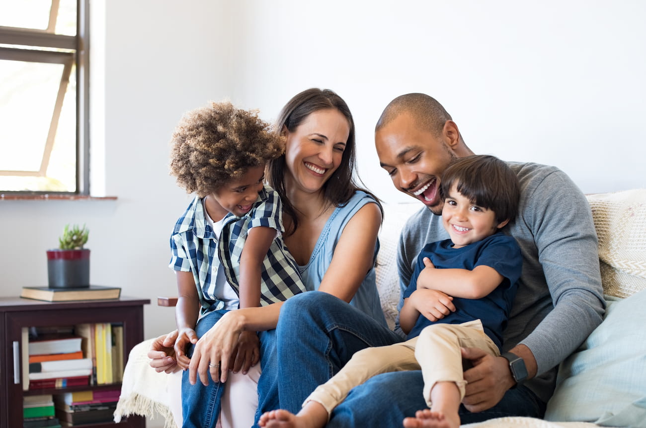 http://A%20smiling%20family%20of%20four%20sits%20on%20a%20couch,%20with%20two%20children%20on%20the%20parents'%20laps.%20A%20window%20and%20small%20table%20are%20in%20the%20background.