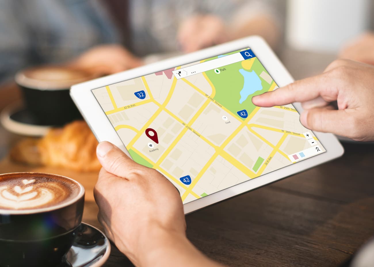 http://A%20person%20uses%20a%20tablet%20displaying%20a%20digital%20map%20with%20a%20location%20pin,%20while%20a%20coffee%20cup%20and%20pastries%20are%20on%20the%20table.