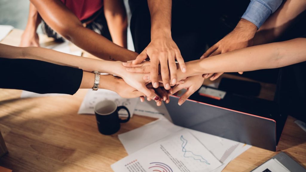 Hands of multiple people stacked in the center over a wooden table with scattered papers and a mug nearby.