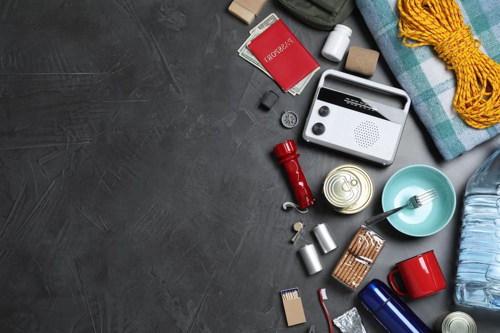 http://Emergency%20preparedness%20kit%20materials,%20such%20as%20flashlight,%20canned%20food,%20batteries,%20radio,%20and%20blankets,%20scattered%20on%20a%20grey%20floor
