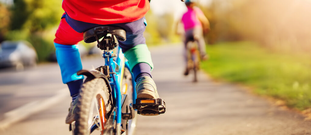 Two people riding bicycles along a paved pathway with greenery in the background.