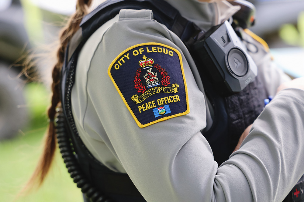 Close-up of a uniformed municipal peace officer’s shoulder and torso, showing a “City of Leduc Enforcement Services – Peace Officer” shoulder patch and a body-worn camera mounted on the chest.
