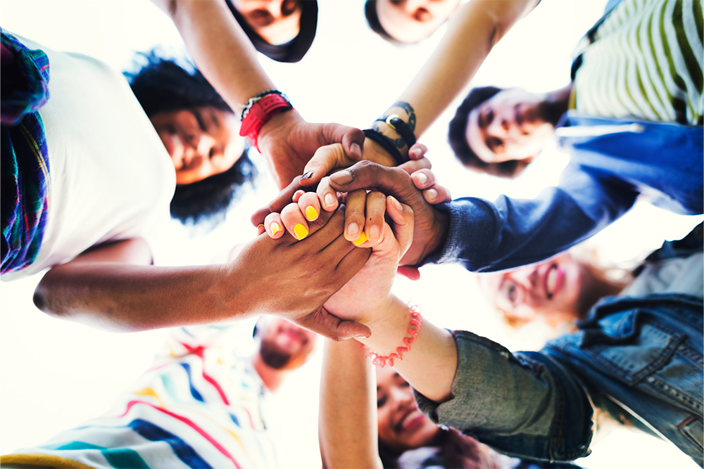 Group of young people standing in a circle with hands stacked together in the center, symbolizing teamwork and community connection.